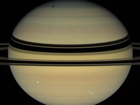 Saturn's rings cast dark bands across cloud tops in the northern hemisphere. Near the pole, an elongated shadow can be seen from Saturn's moon Tethys. Icy moons Dione (front right) and Enceladus (back right) are also seen by NASA's Cassini spacecraft.