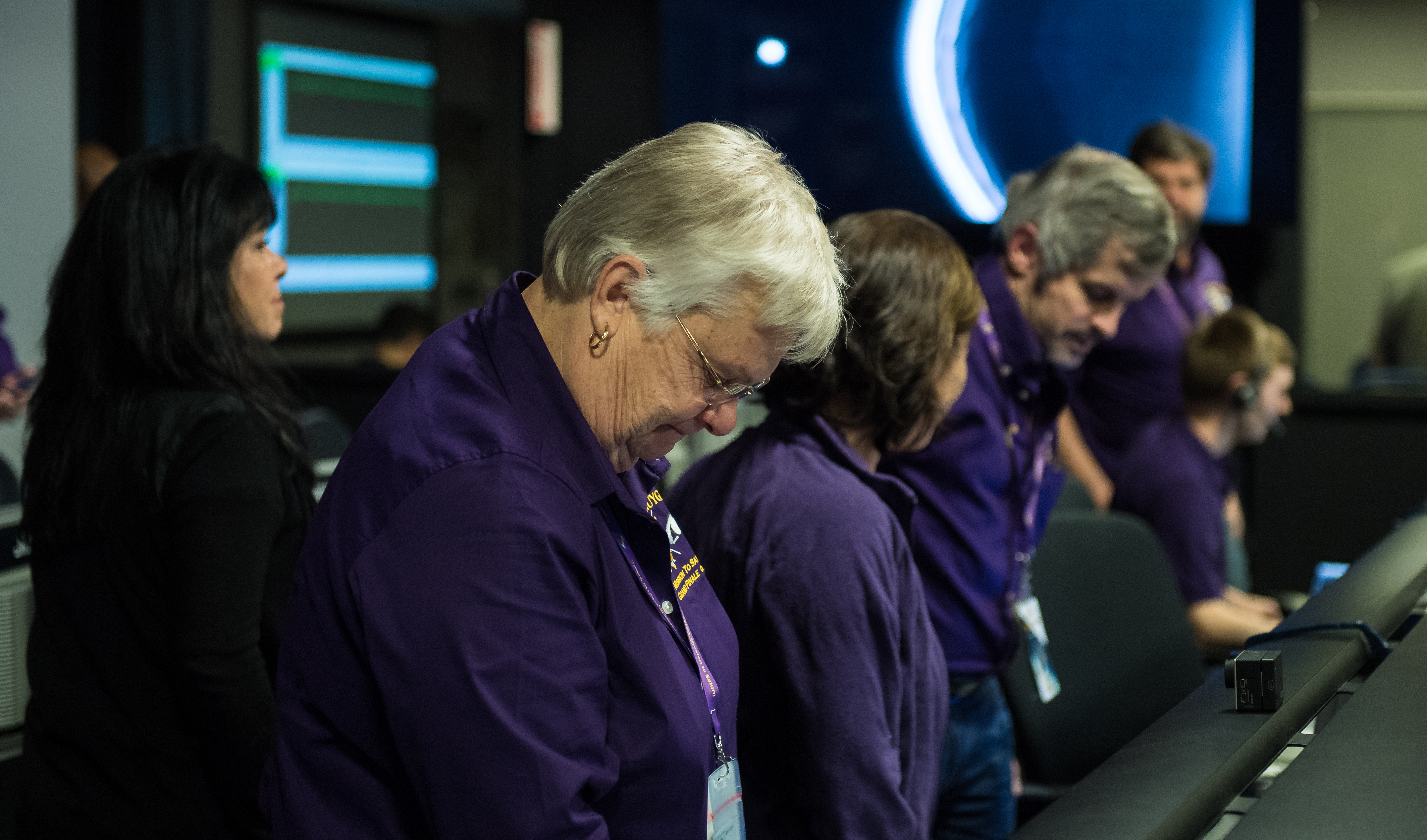 Color image of people in mission control with their heads bowed.