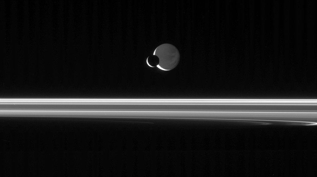 The Cassini spacecraft looks across the unlit ringplane as Mimas glides silently in front of Dione.