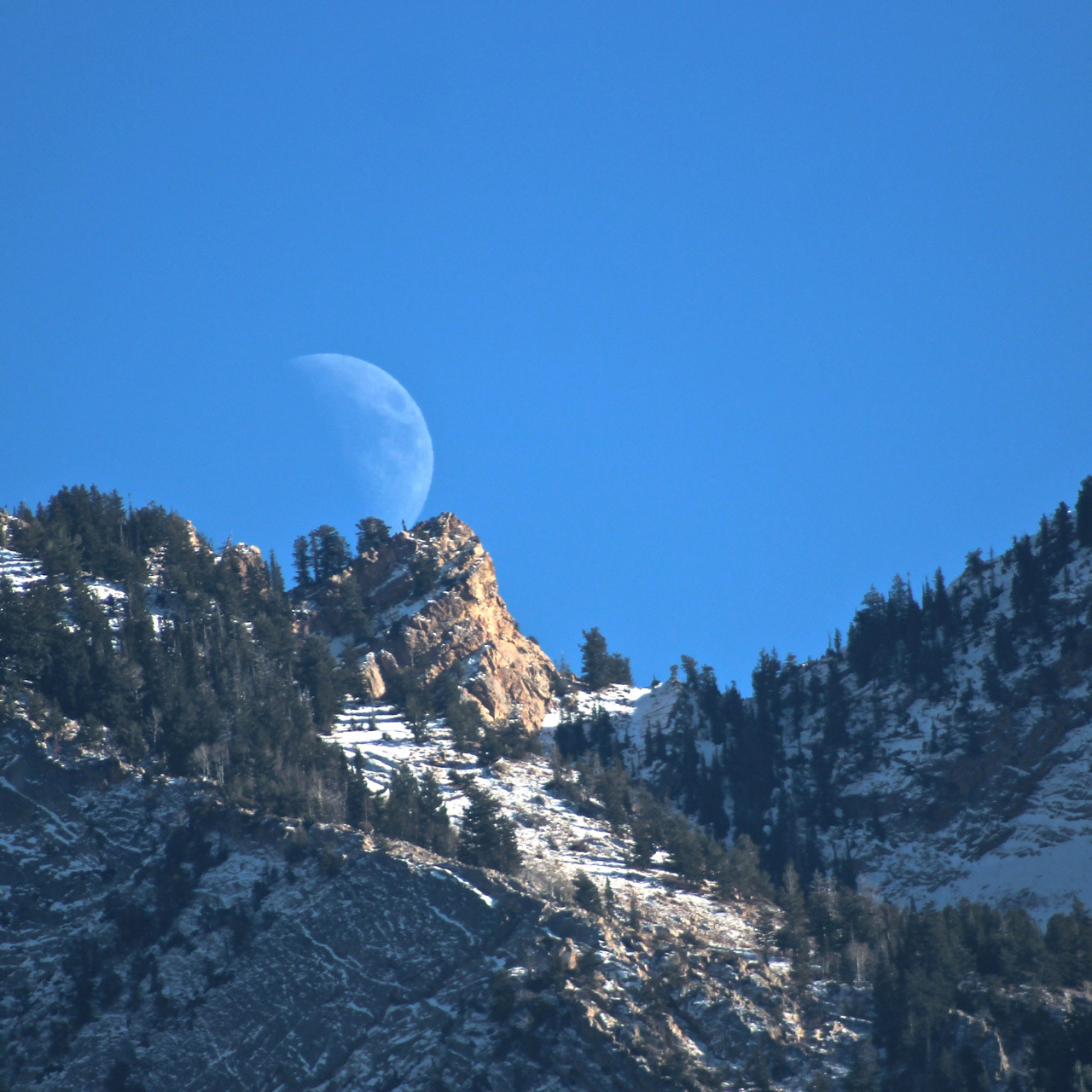 Moon rising over a mountain ridge in daylight.