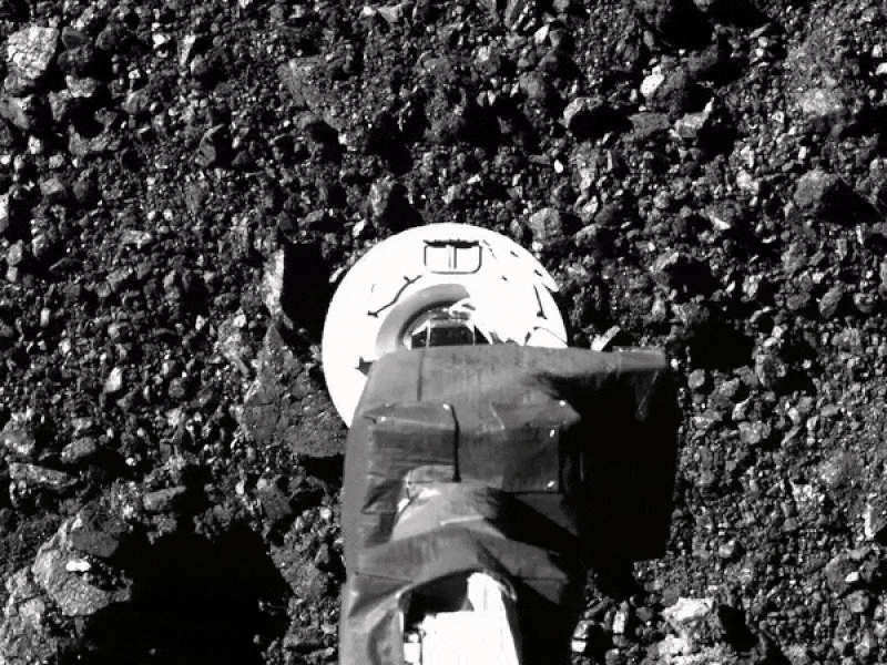 In this black and white animated image, a circular device stretched out from a robotic arm descends quickly toward a rocky surface, touches it, and then ascends as debris flies all around.