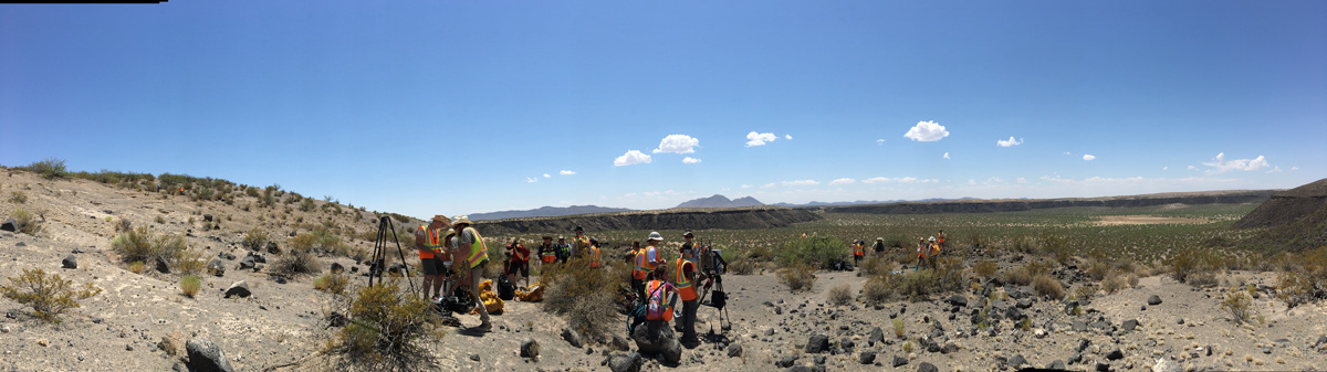 Panoramic image of several researchers wearing orange vests, all at work in a scrubby desert landscape. Mountains and mesas are visible in the background.