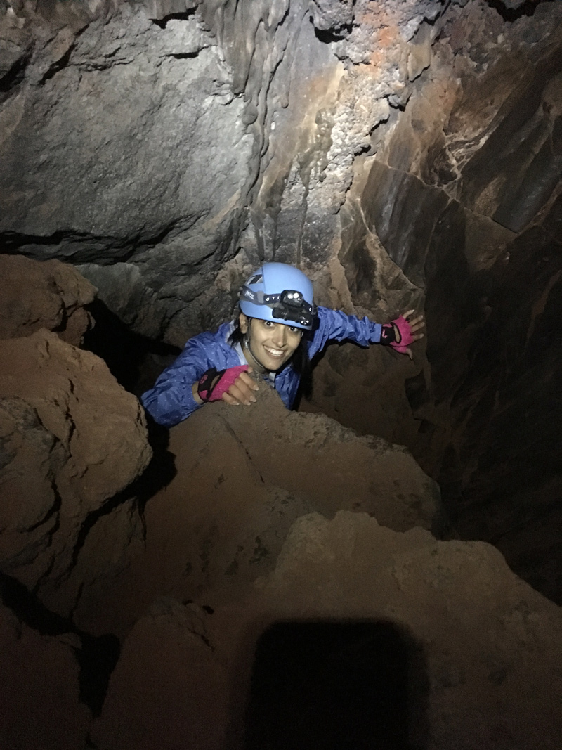 A smiling researcher emerges from a narrow passage in an underground cave. She wears a helmet and headlamp. 