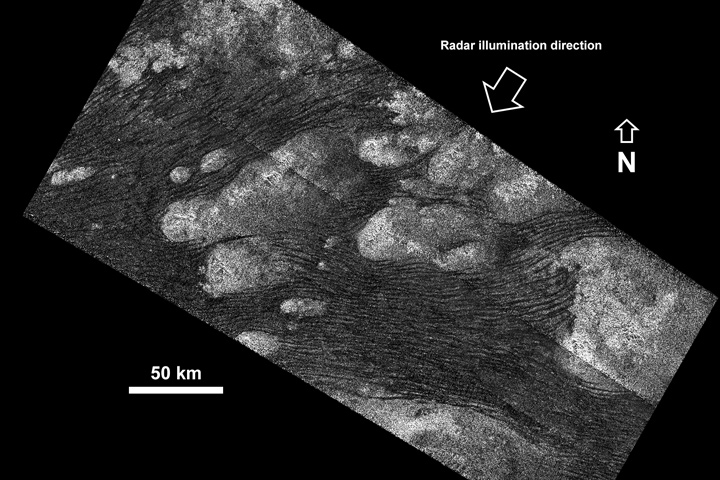 Black-and-white radar image of a ripplng field of sand dunes interrupted by light-colored patches. A 50 km scale bar shows that this image represents a larger area, with larger features, than its companion.