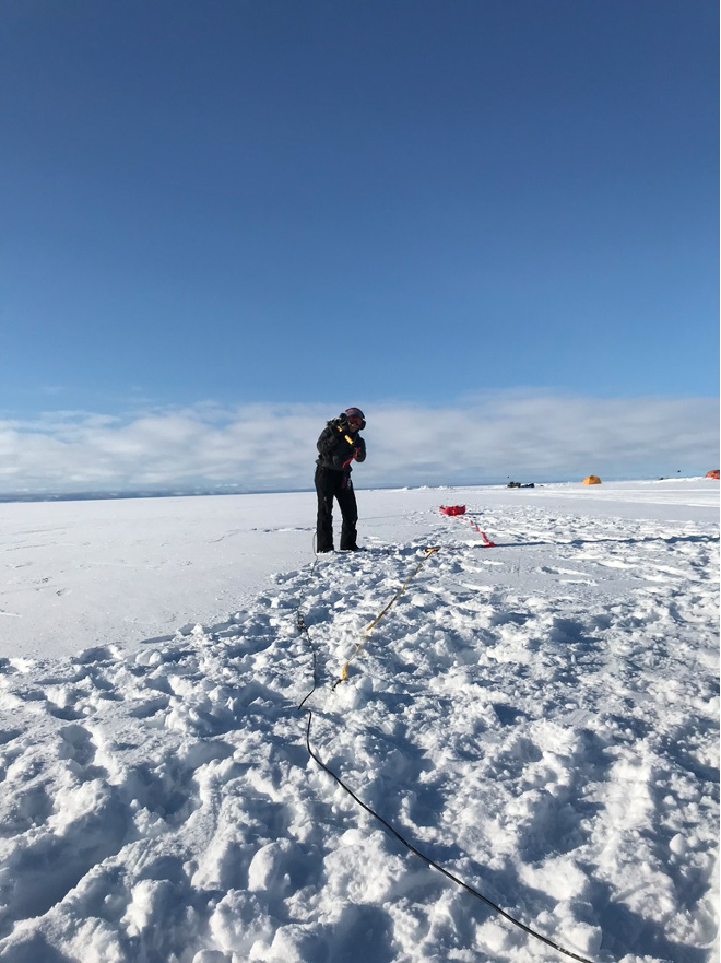 On a field of snow beneath a blue sky, a researcher raises a hammer over her shoulder.