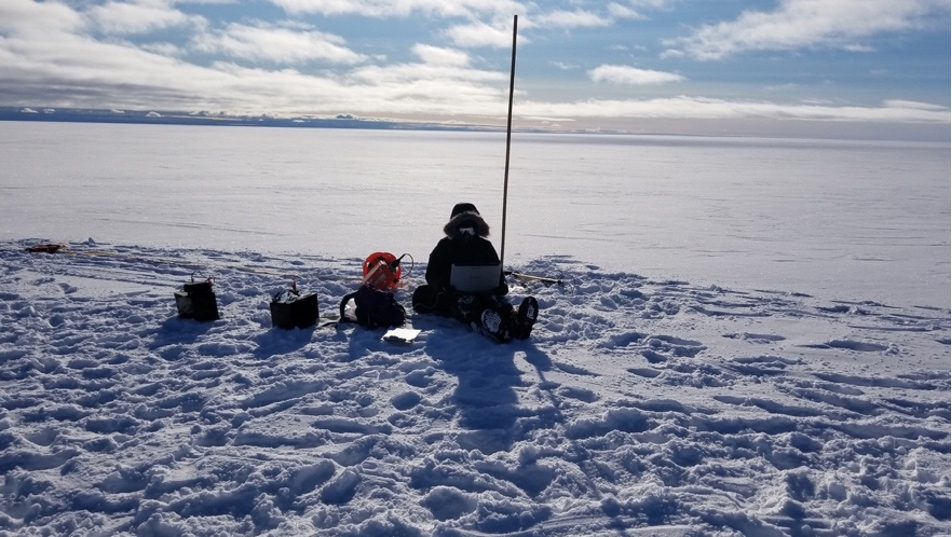 A researcher sits in the snow, bundled in protective gear, surrounded by small pieces of equipment.