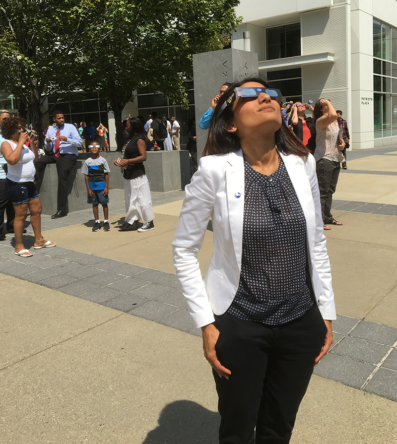 A woman wearing eclipse glasses stands outside and looks up at the sky. Several people, some also wearing eclipse glasses and looking upward, appear in the background.
