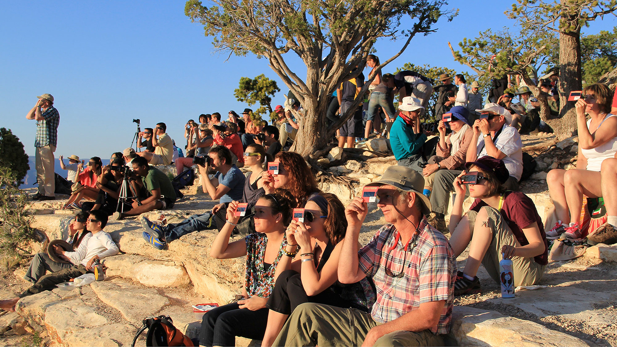 A crowd of people sitting on a rocky slope with a few trees, all looking in the same direction and wearing either solar viewing glasses or holding up handheld solar viewers.