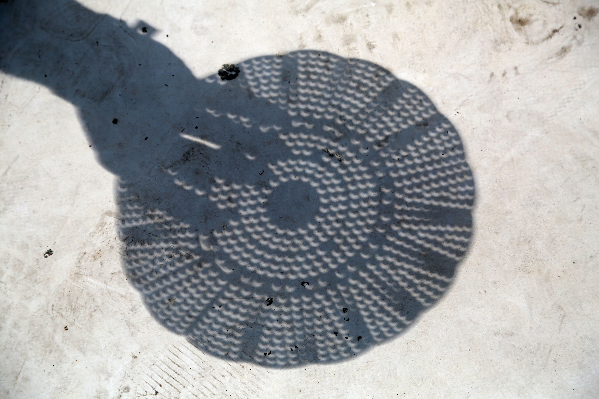 The shadow of of a colander and a hand holding it, with the circular hole creating crescents on the ground during an eclipse.