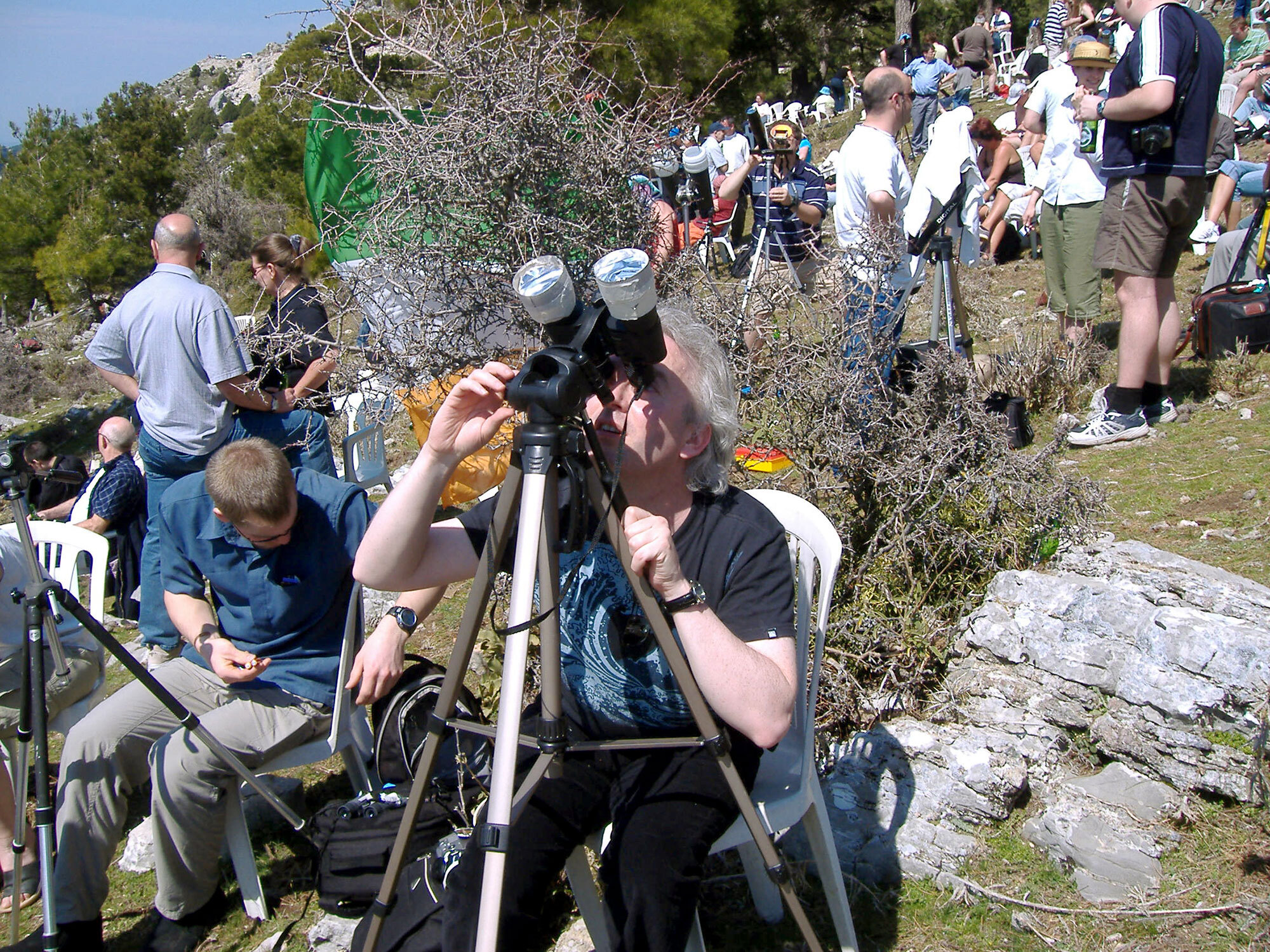 In the foreground, a woman sits in a chair and looks through a pair of binoculars affixed on a tripod and pointed upward. The binoculars have silver-colored coverings over the two ends of the binoculars that are pointed upward toward the Sun. Other people are seen sitting and standing around the woman and in the background.