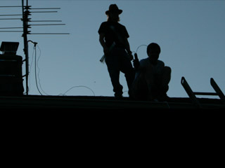 Silhouettes of two men on a rooftop with a radio antenna to their left.