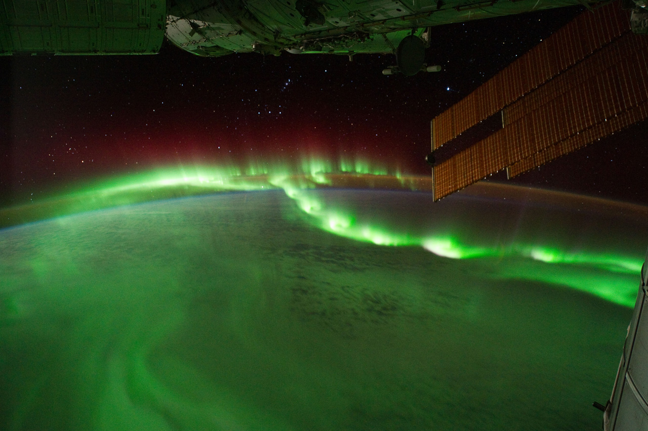 An image taken from the International Space Station (ISS) of auroral beads dancing in Earth’s atmosphere, bright green glowing bands of light. The atmosphere can be seen as a thin glowing orange line above Earth. Lower atmosphere clouds can be seen below the aurora. Parts of the ISS can be seen on the top of the image. Solar panels jet out from the top left corner. Above the Earth is space dotted with stars.