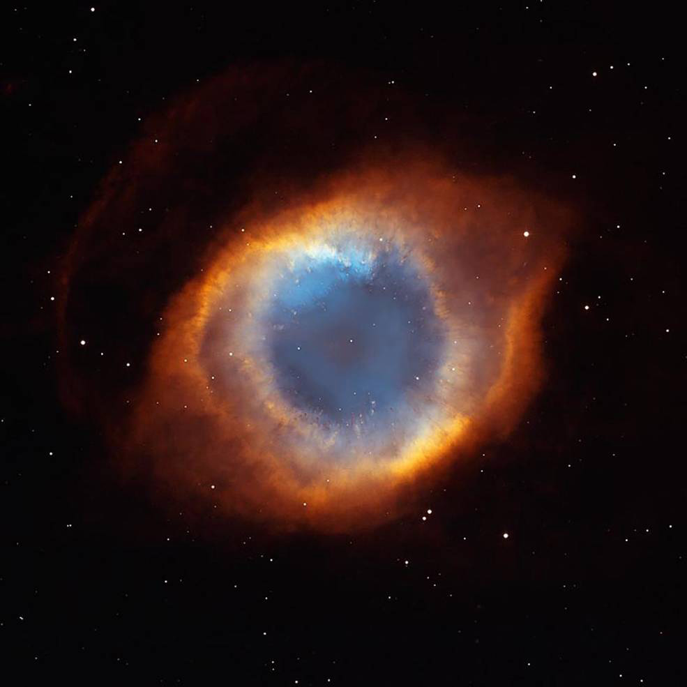 An image of the Helix Nebula, taken by the Hubble Space Telescope. This tunnel of glowing gasses looks like an eye floating in space. A blue cloudy pupil at the center, surrounded by an orange -yellow iris.White stars dot the entire image. 