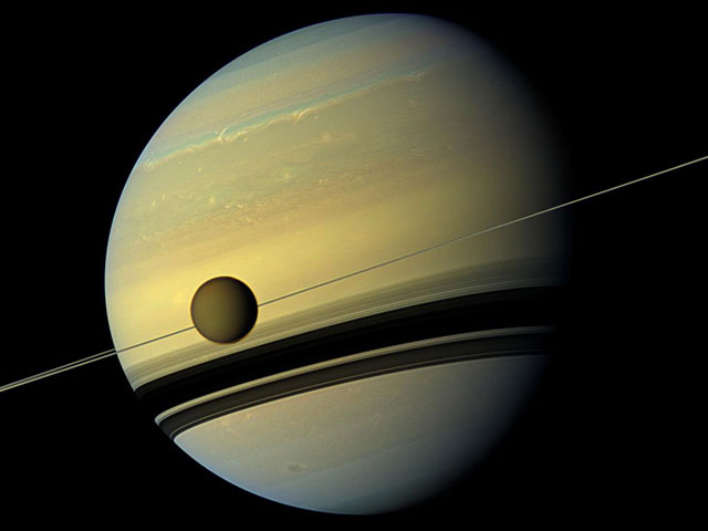 The large moon Titan looms in front of Saturn in this image taken from orbit. Saturn&#39;s rings are edge-on appear thin, but their shadow on the planet reveal their true size.