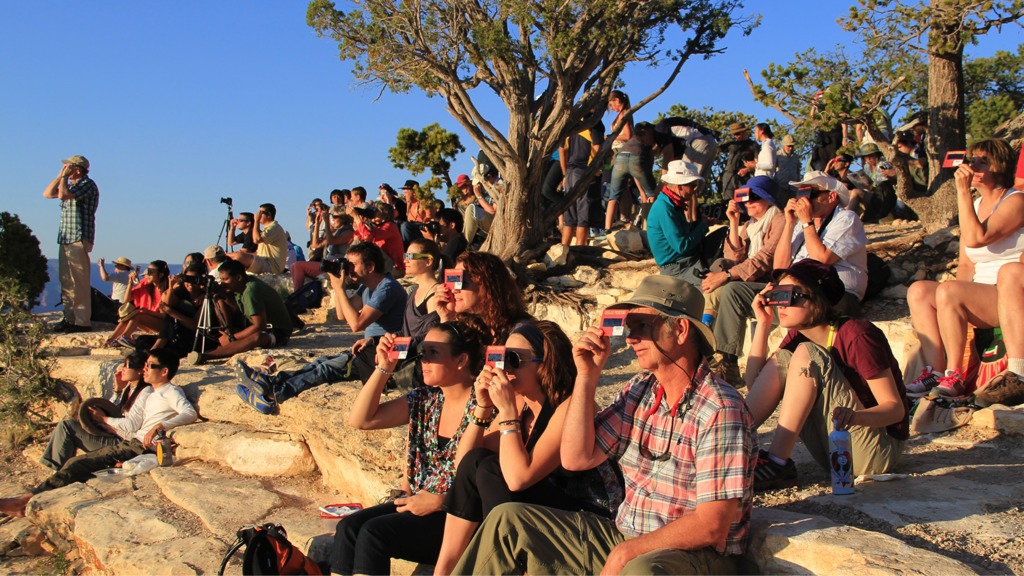 A large group of people sit on rocks, all looking in the same direction. They hold red cards with solar filters in front of their eyes.