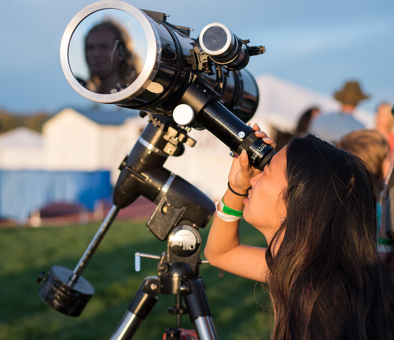 A person looks through a telescope during the daytime. A solar filter is attached to the front of the telescope.