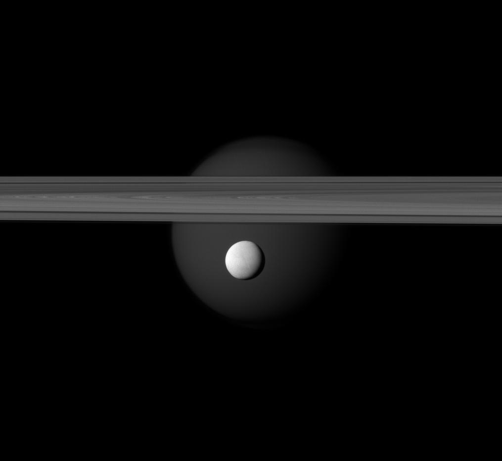 The brightly reflective moon Enceladus appears before Saturn's rings while the larger moon Titan looms in the distance.