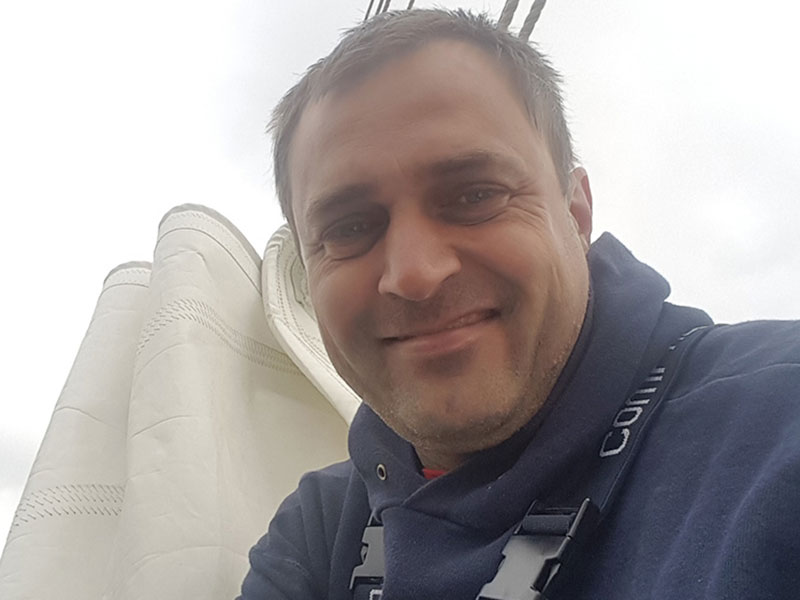 A close up of Peter on a sailboat with white sails furled in the background.