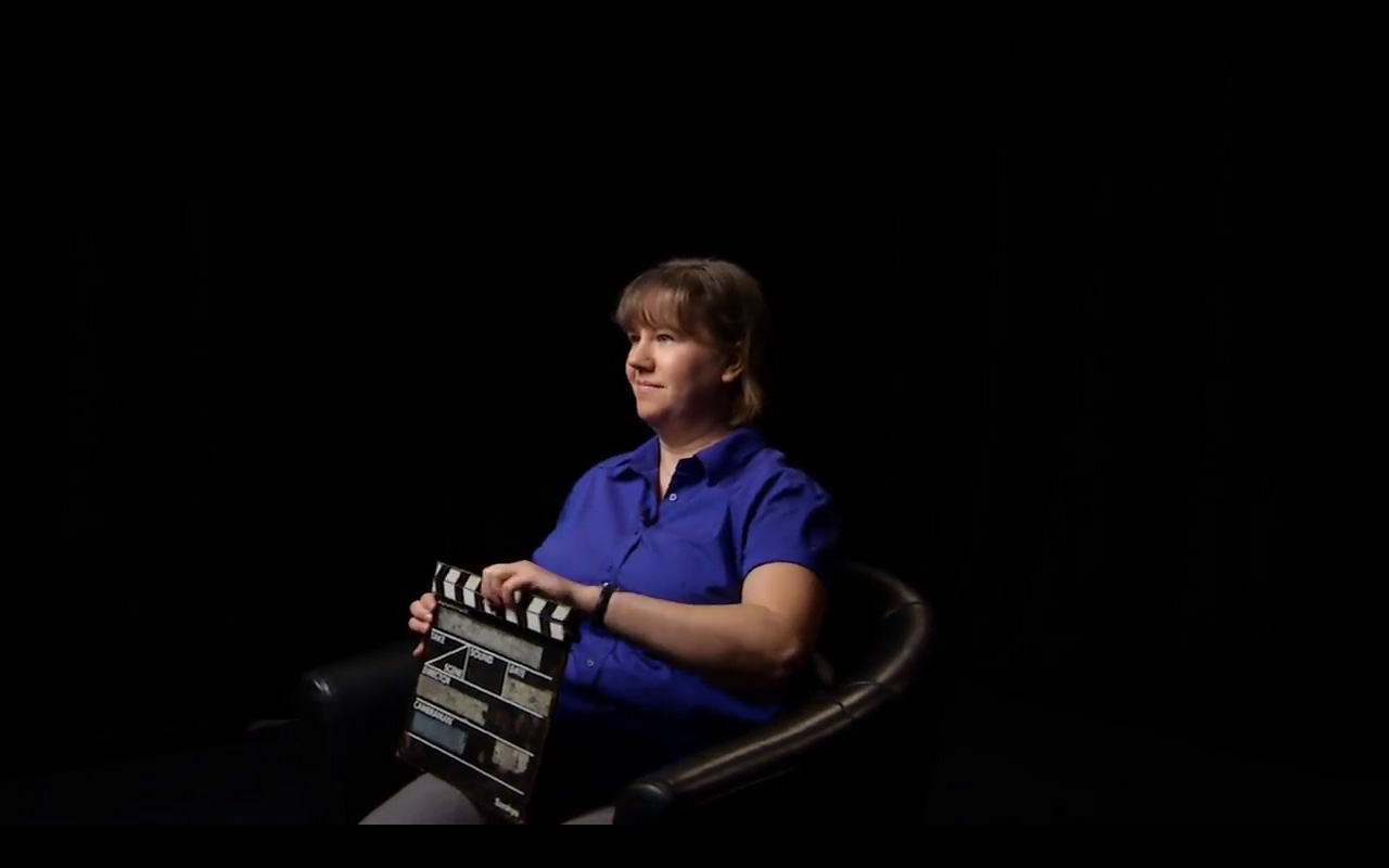 Sarah sitting in a director's chair on a dark set.