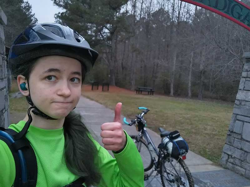 Emily, wearing a bike helmet, is giving a thumbs up from the edge of a woodland biking trail.