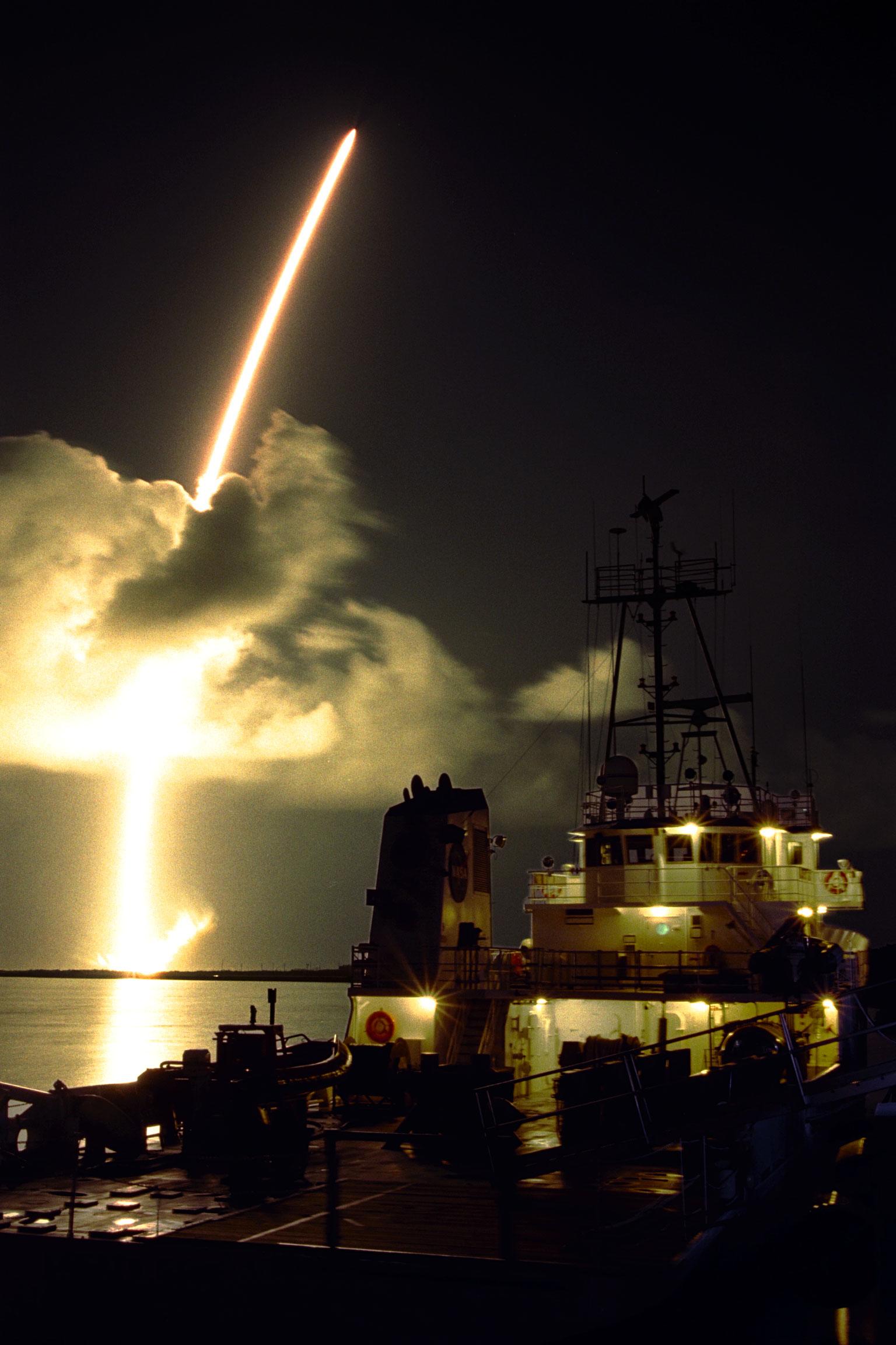 Color image of a rocket launch with a ship in the foreground.