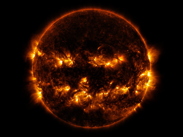 A dark red circular sun with bursts of bright orange light that create something like a jack-o-lantern’s face 