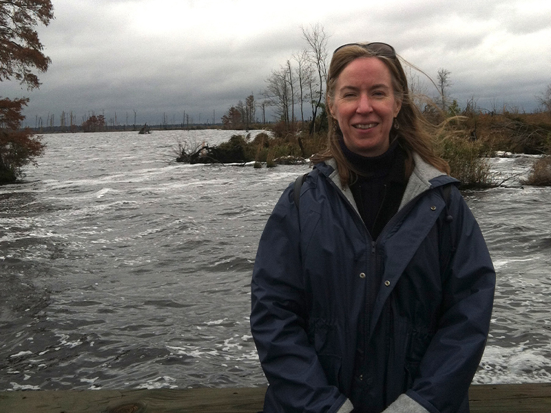 Lin Chambers wears a blue coat with gray lining as she poses for a photo in front of foamy water with islands of trees under a cloudy, gray sky. 