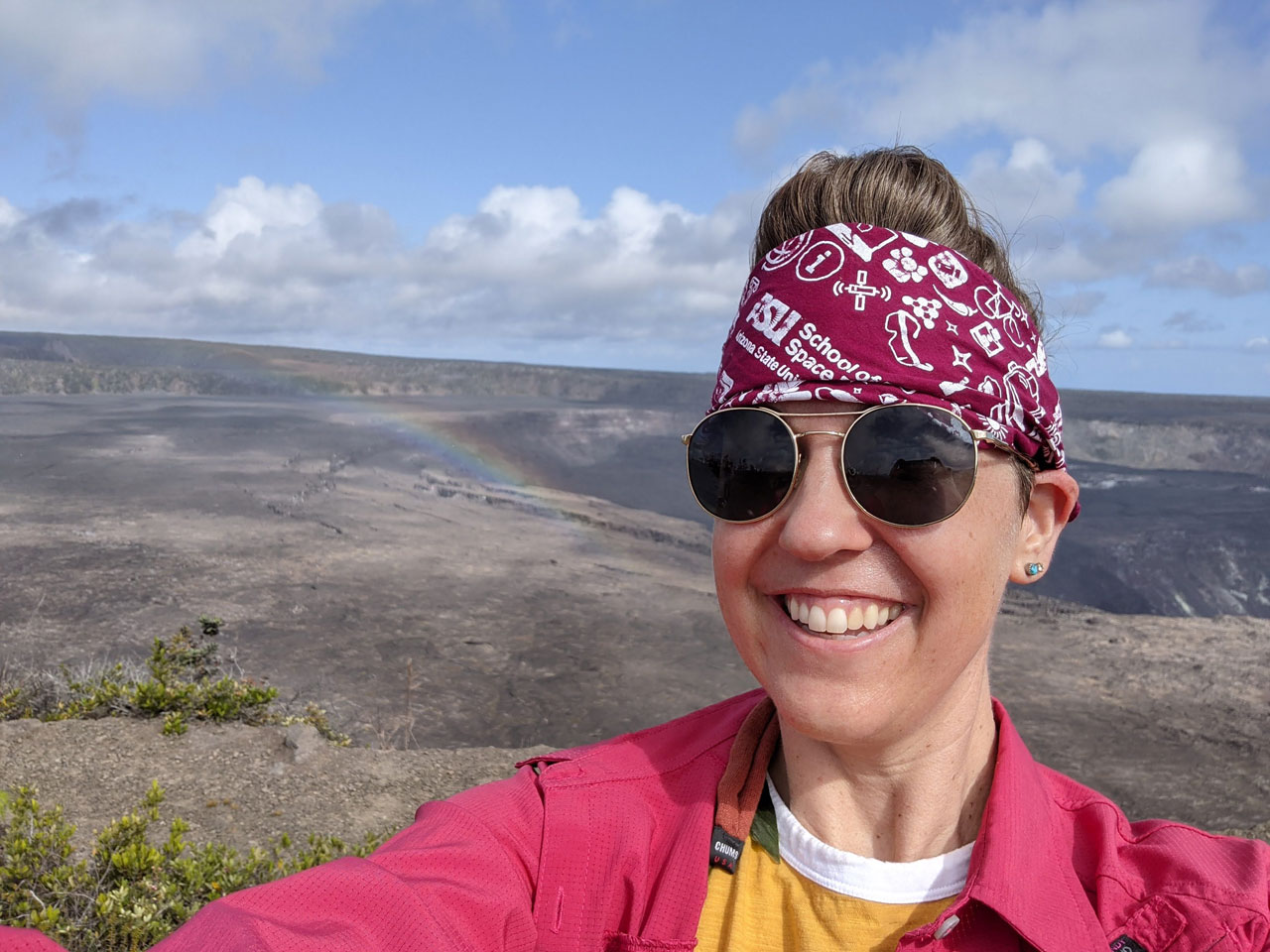 Melanie is taking a selfie in front of a lava field. She's wearing dark aviator glasses and a red jacket.