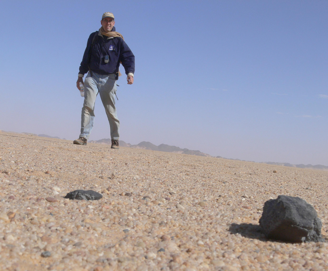 Man standing over small meteorite in desert sand.