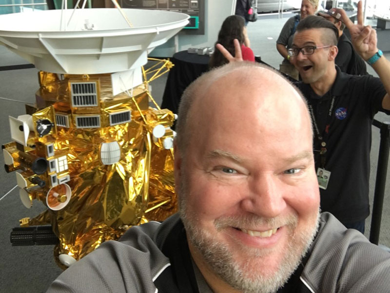 Phil is wearing a NASA shirt. His co-worker is making V for victory signs in the background.
