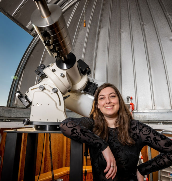 Ciera has her elbow propped on the telescope platform. She is standing in an observatory dome and wearing a black floral print shirt.