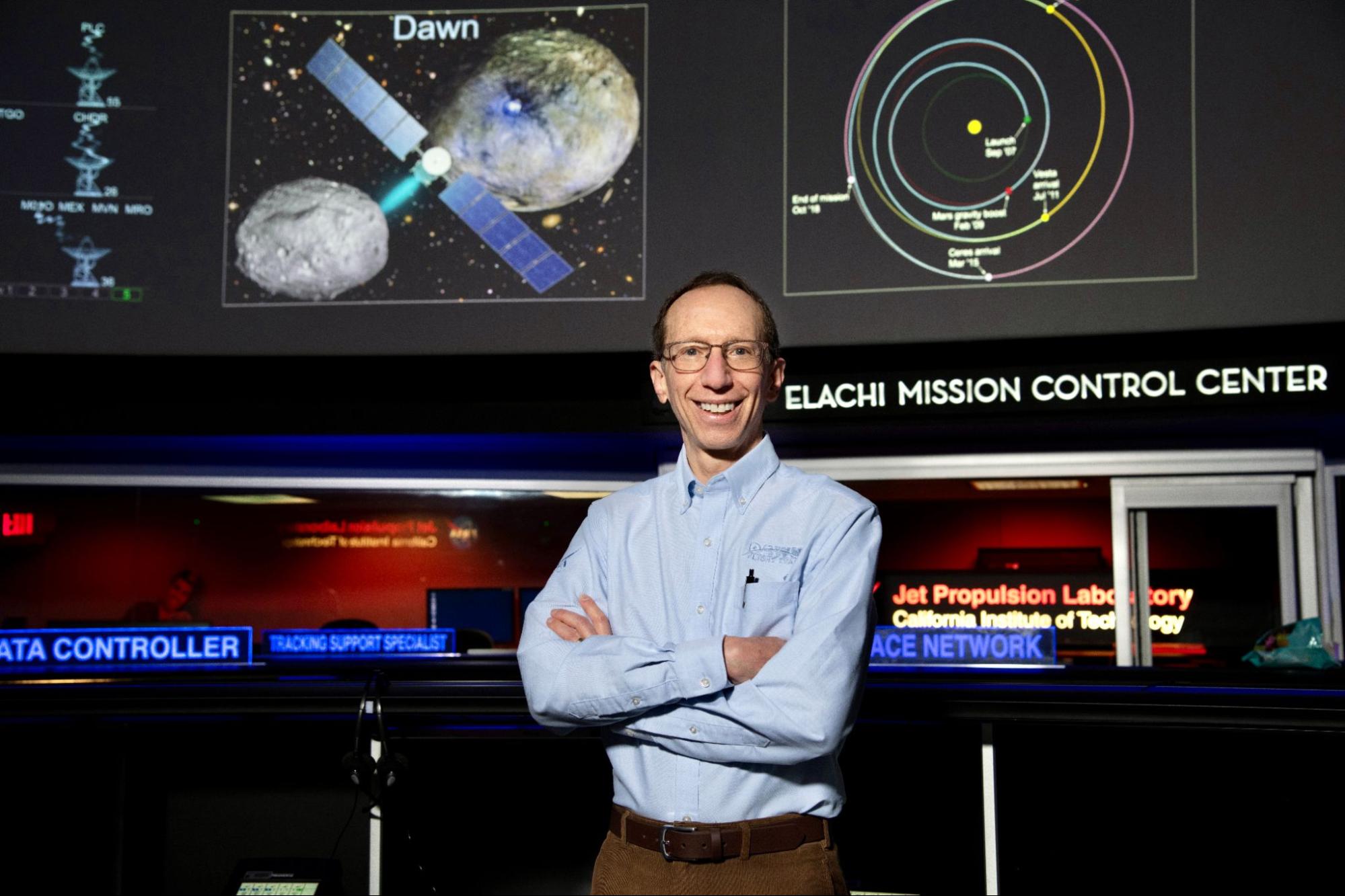 Marc Rayman smiling and standing with his arms crossed in Mission Control with an illustration of the Dawn spacecraft, its two destinations, and the flight path on the wall behind him. 