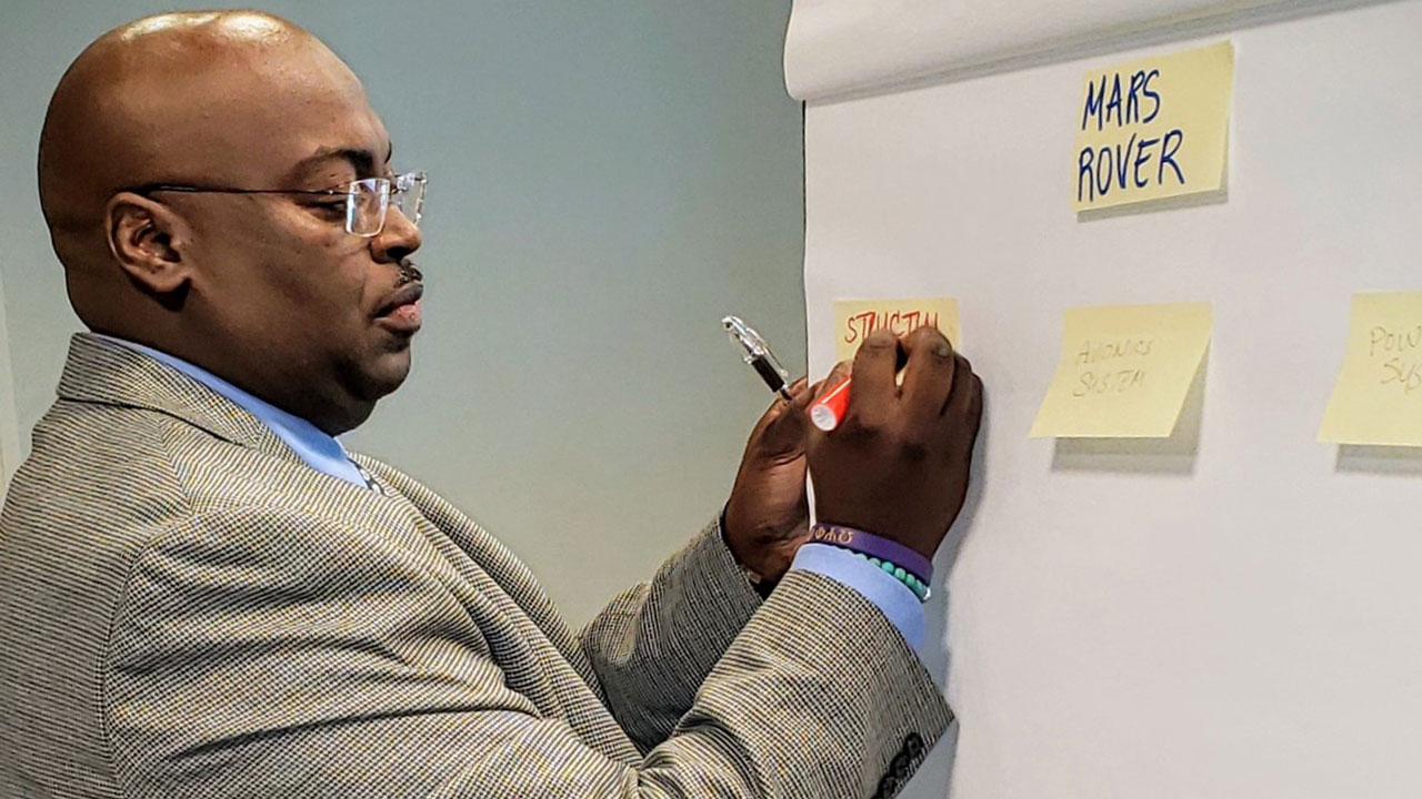 Clarence is standing at a whiteboard writing on yellow sticky notes.