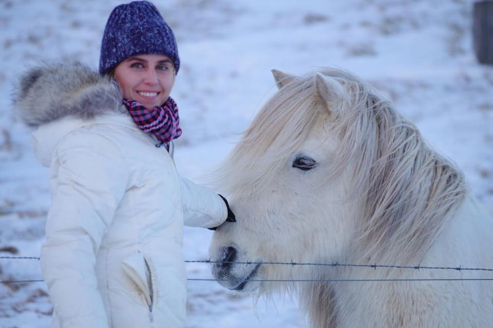 Rachael in the snow petting an Icelandic horse. She's wearing a white parka and the horse has a white coat and mane.