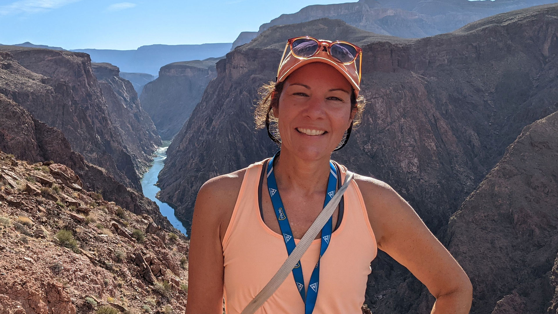 In this photo, Lori is standing in front of the steep, rugged chasm of the Grand Canyon, with the blue Colorado River below. She's wearing light hiking clothes.