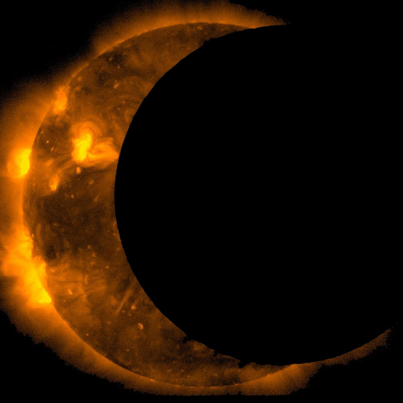 Image of a partial solar eclipse, with the Sun being blocked by the Moon. About 25% of the sun is blocked, making it appear as a crescent with sunspots and prominences visible on the surface and in the corona. The Moon is black and in the foreground of the image.