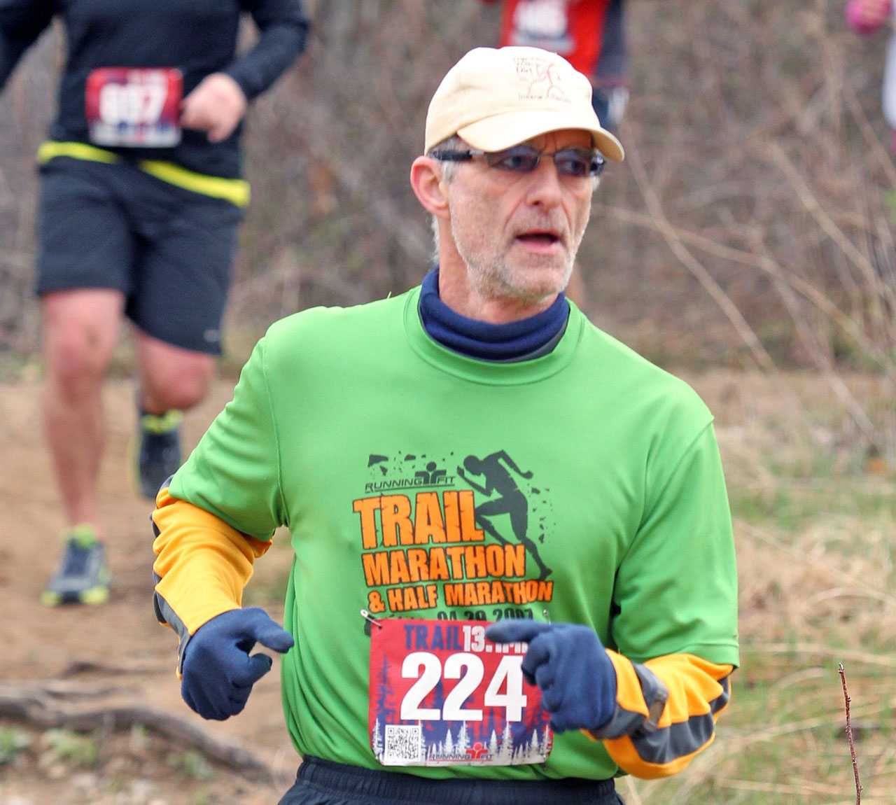 Michael is running on a dirt trail. He's dressed for warm weather with gloves and a thermal undershirt over a green race T-shirt.
