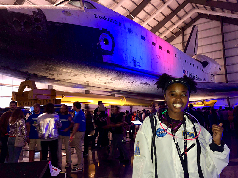 Janelle Wellons in a NASA jacket standing in front of a Space Shuttle illuminated with colorful party lights.