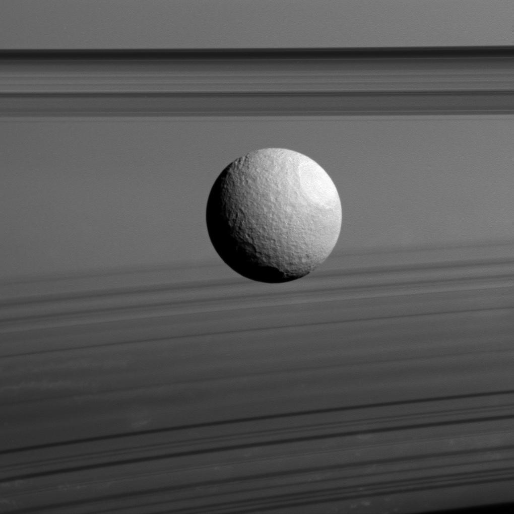 Black and white image of Saturn's moon Tethys in front of Saturn's rings.