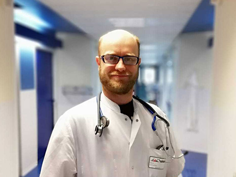 Guillaume stands in a hospital hallway. He is wearing a lab coat and a stethoscope around his neck.