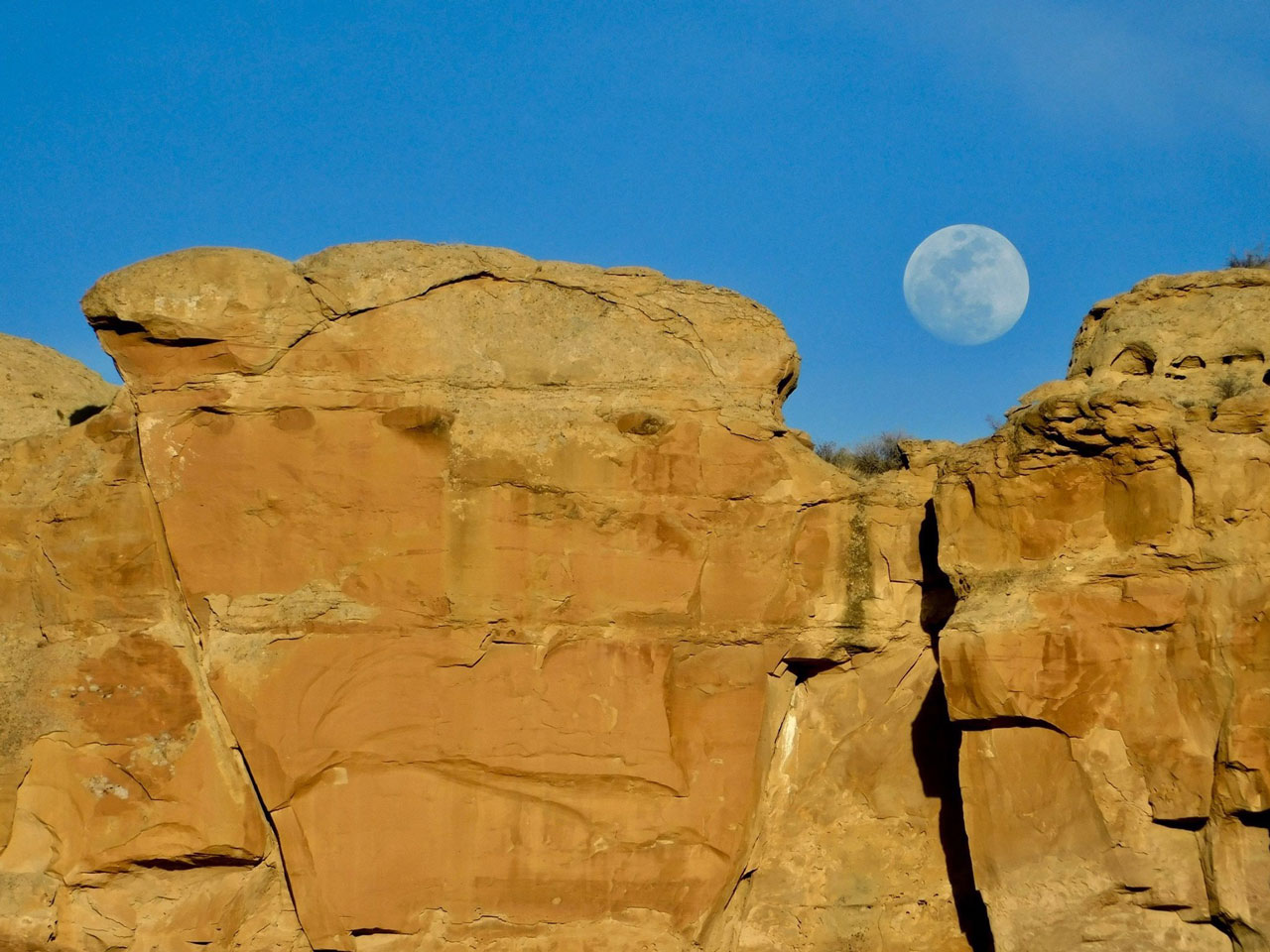 The full Moon rises in daylight above ancient ruins carved into a steep desert cliff face.