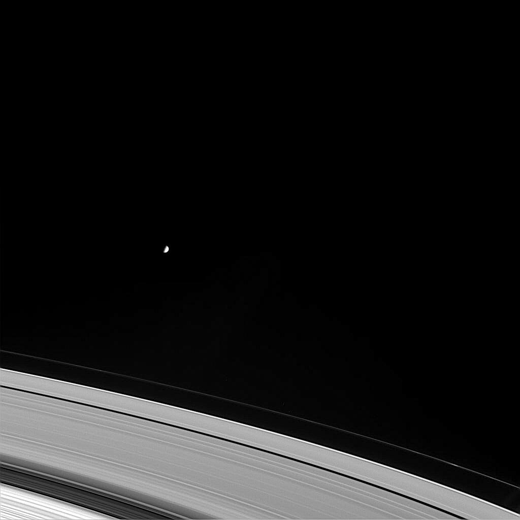 Black and white image of Mimas as a small white dot above the expanse of Saturn's rings.
