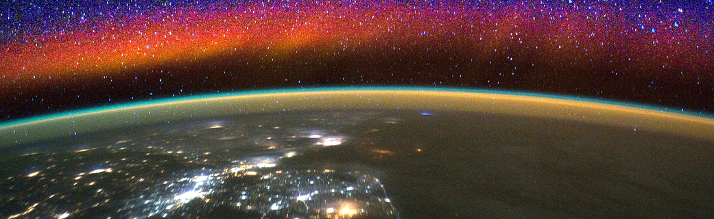 A portion of the curve of the Earth, seen from space. Earth twinkles with lights at night. Separating Earth from the starry sky are bands of yellow and orange, the atmosphere. Above that are clouds of red and orange with stars shining through.
