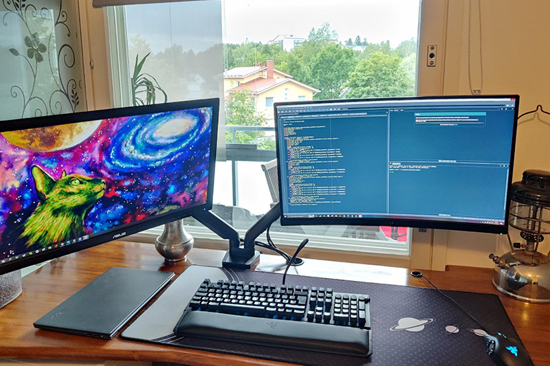 Two computer monitors on a swingarm sitting on a desk with a keyboard and mouse with a view out a window.