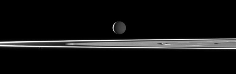Two light sources illuminate Saturn's highly reflective moon Enceladus in this image featuring bright rings. Most of the moon is dimly lit by sunlight reflected off Saturn. However, a thin crescent of the moon is lit by sunlight on the moon's leading hemisphere.