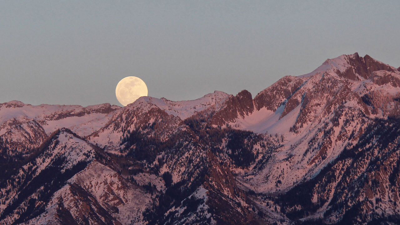A full Moon rises above rocky, snow-capped mountain peaks.