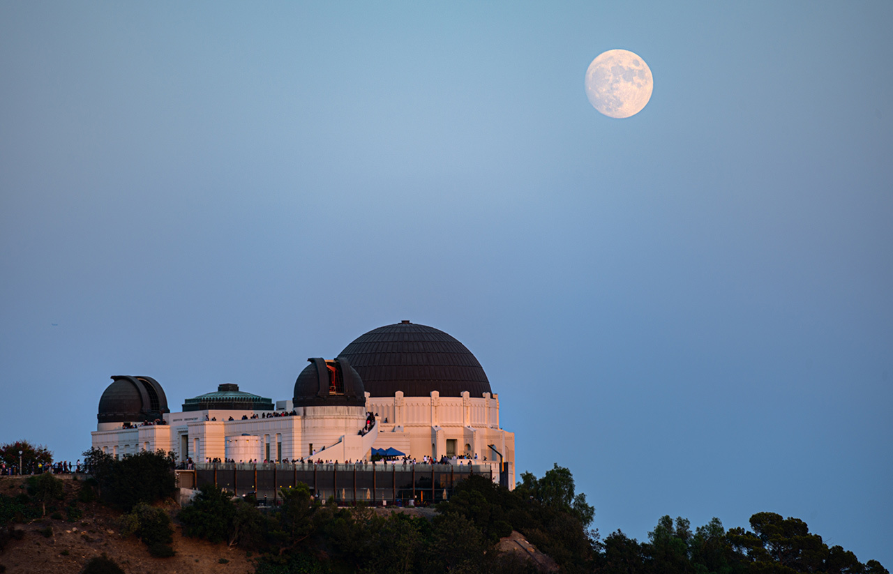 Moon rising over a white building with domes sitting on a hillside.