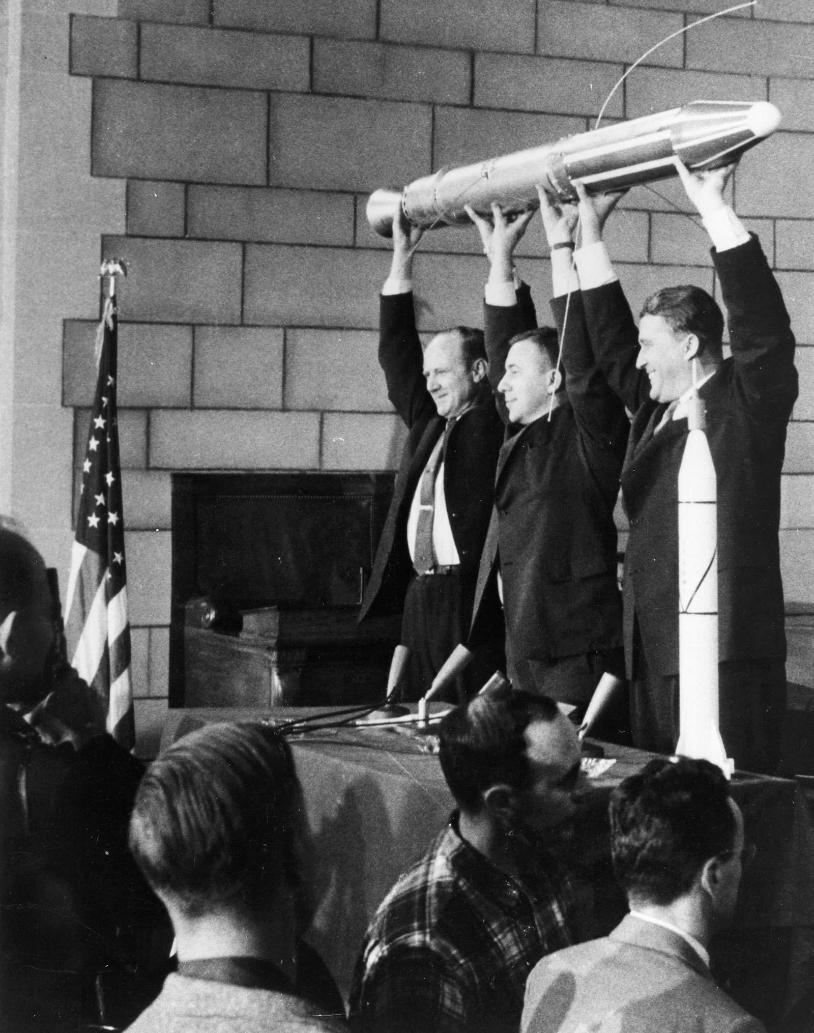 Three smiling men holding spacecraft model above their heads.