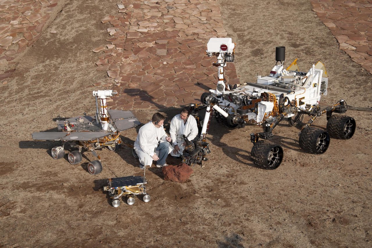 Two spacecraft engineers crouch next to rovers.