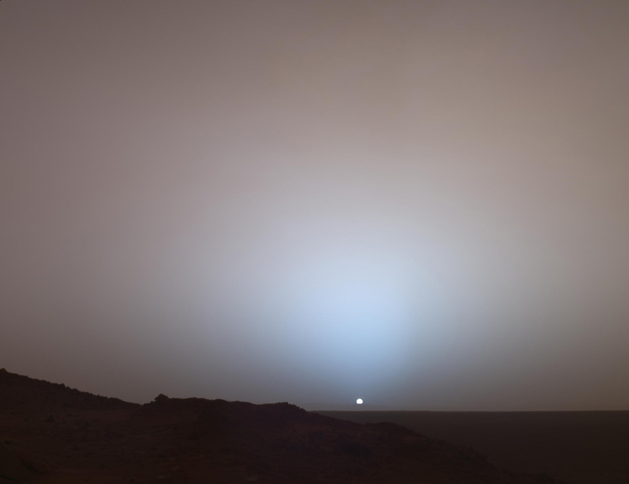 Tiny sun rises on the horizon of a Martian plain. A robotic arm is visible in the foreground.